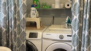 A laundry area with two washing machines, a shelf with cleaning supplies, and patterned curtains.