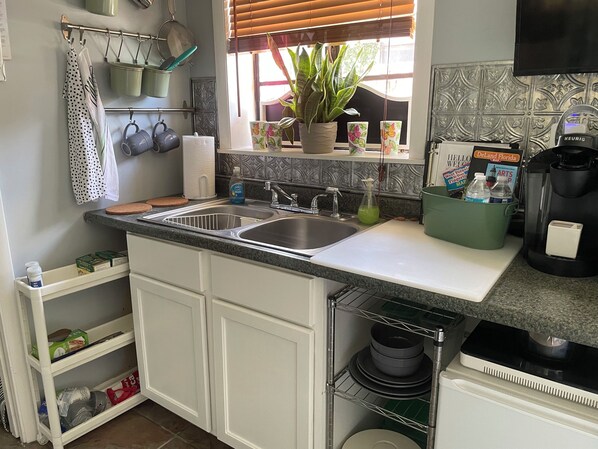 Une cuisine Ă©quipĂ©e dâun Ă©vier Ă deux bacs, dâune machine Ă cafĂ©, dâune plante en pot et dâune Ă©tagĂšre contenant des ustensiles de cuisine.