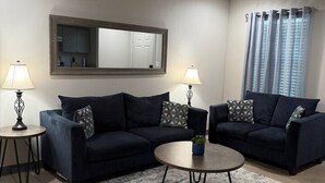 Una sala de estar moderna con dos sofás azul oscuro, una mesa de centro redonda y dos mesitas de noche con lámparas. Un espejo grande cuelga de la pared, y se ve una ventana con cortinas.