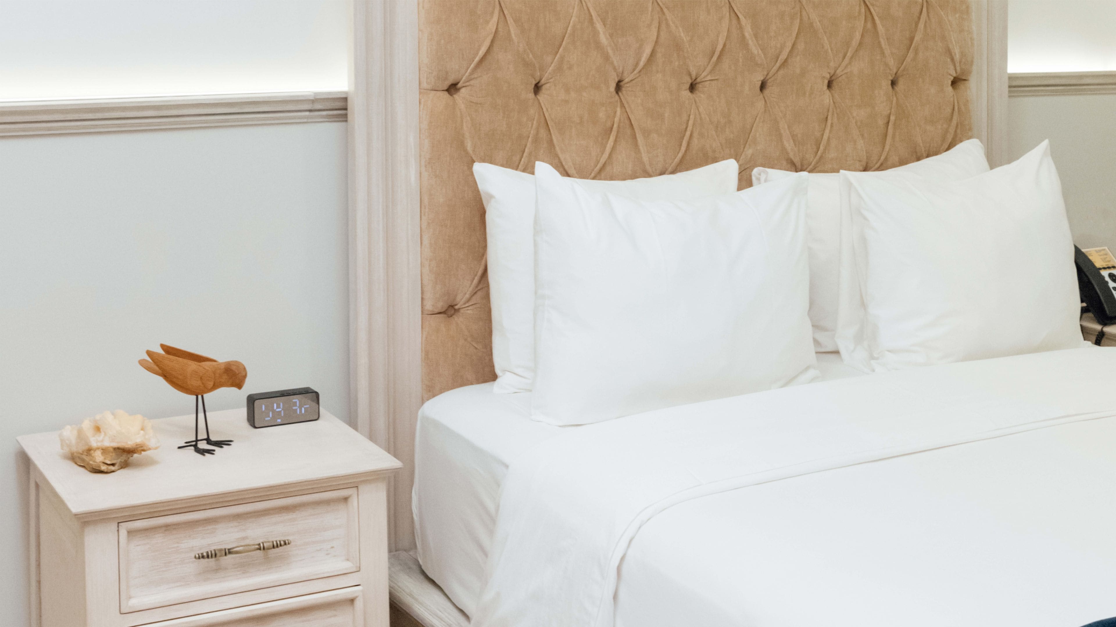 A neatly made bed with white bedding, a wooden nightstand with a clock, and a decorative bird sculpture.