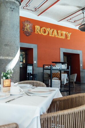 Interior de un restaurante con un letrero que dice 'ROYALTY', mesas con manteles blancos, sillas de mimbre y un pilar de concreto.