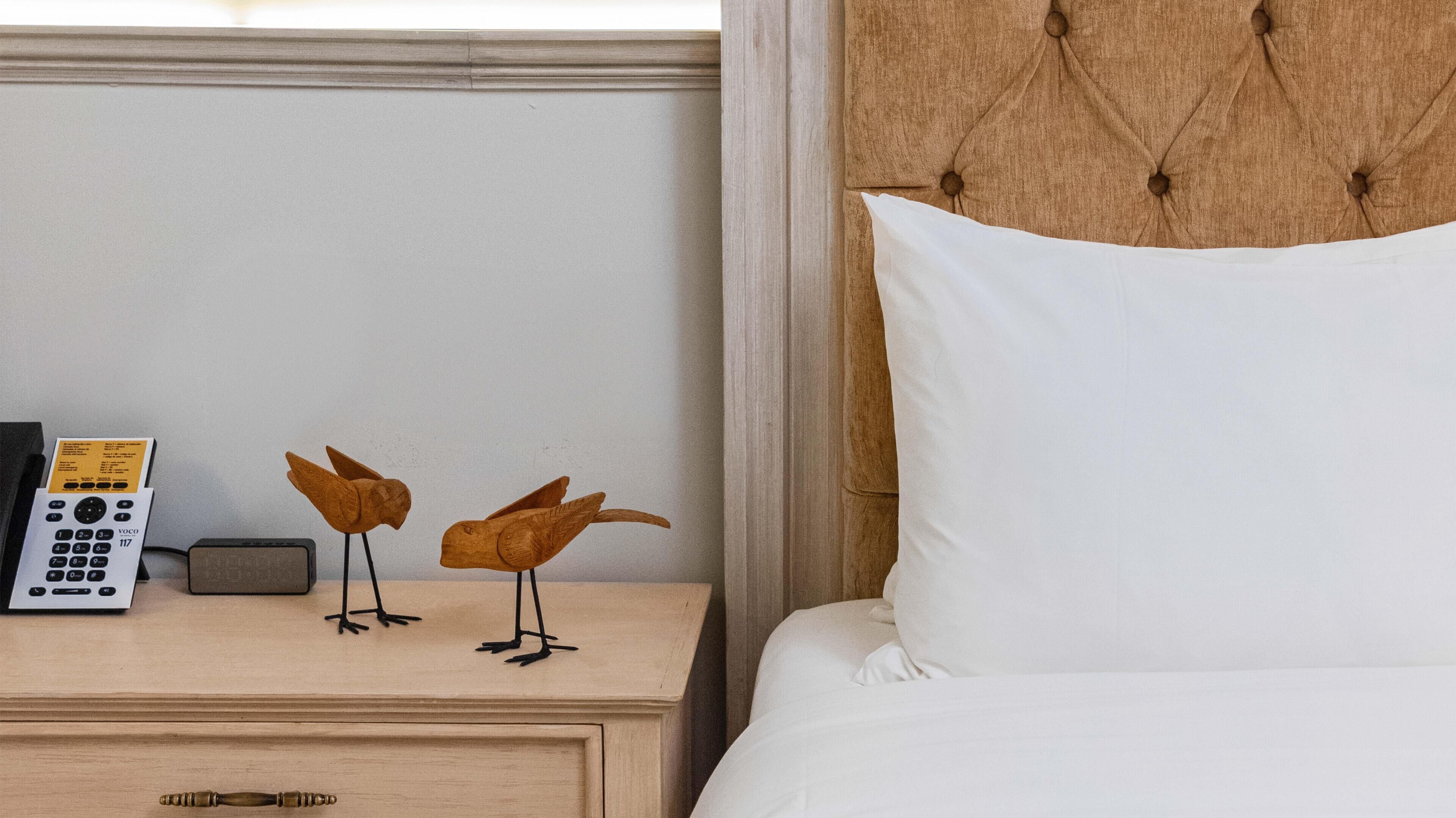 Une chambre d’hôtel avec une table de chevet en bois, un oreiller blanc et deux sculptures d’oiseaux en bois.