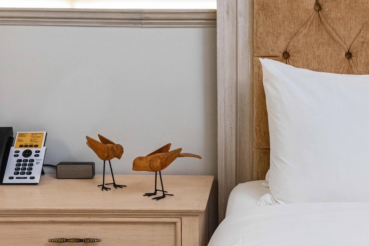 A hotel room with a wooden nightstand, a white pillow, and two wooden bird sculptures.