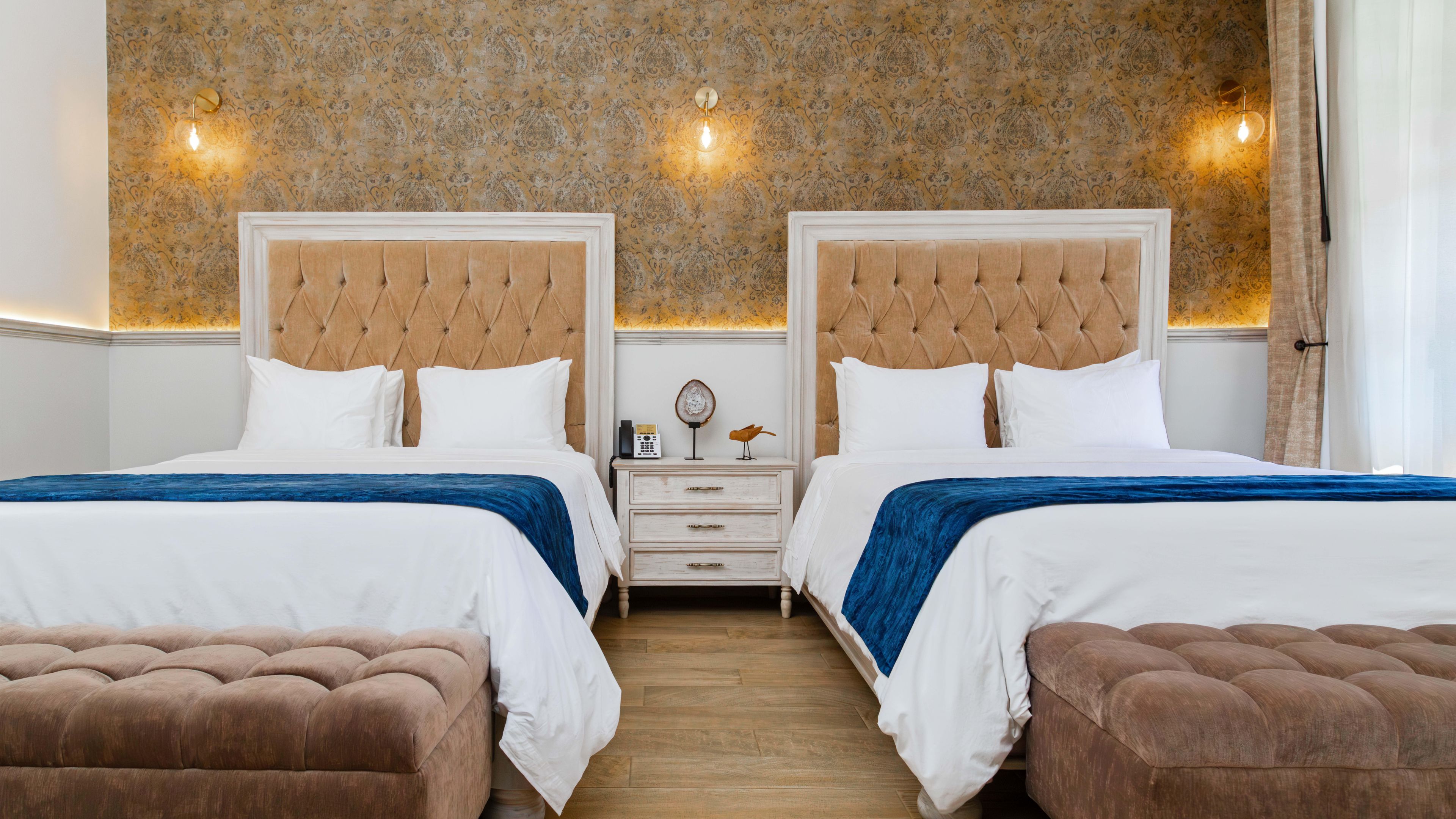 A hotel room with two beds, a nightstand, a wooden floor, and a patterned wallpaper.