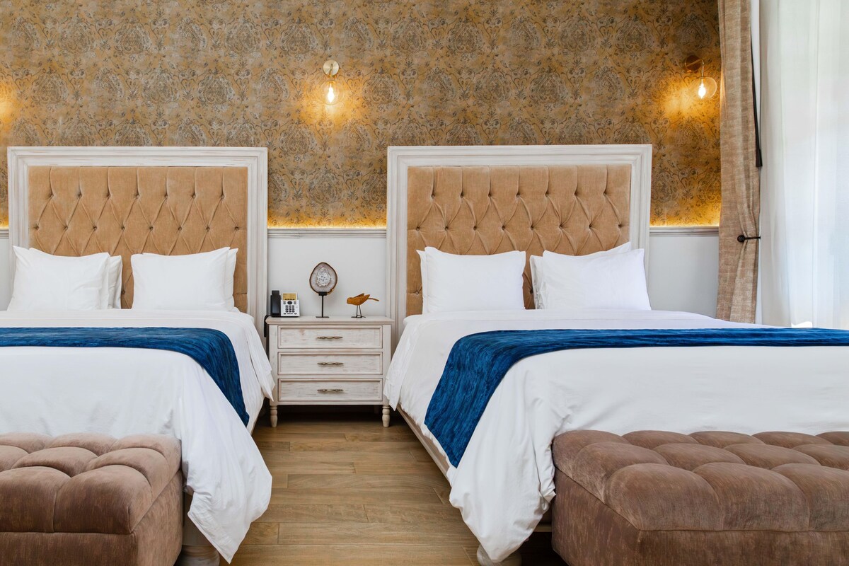 Habitación de hotel con dos camas, una mesita de noche, suelo de madera y papel tapiz estampado.