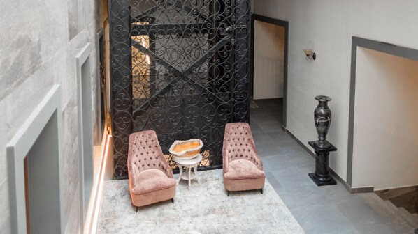 A hallway with a black wrought iron gate, two pink upholstered chairs, a small table with a bowl, and a tall black vase on a pedestal. - Voco Royalty Puebla Downtown (Puebla)