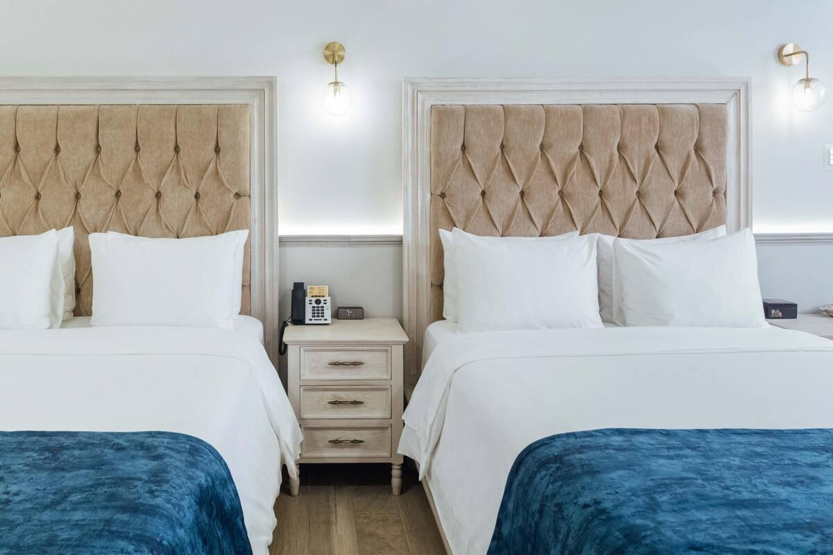 A hotel room with two beds, a nightstand with a phone and a clock, and wall sconces.