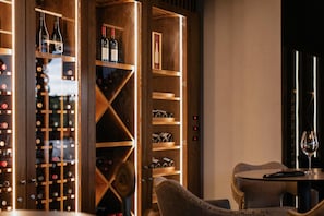 Una bodega con estantes de madera y botellas, una mesa con una copa, y dos sillas.