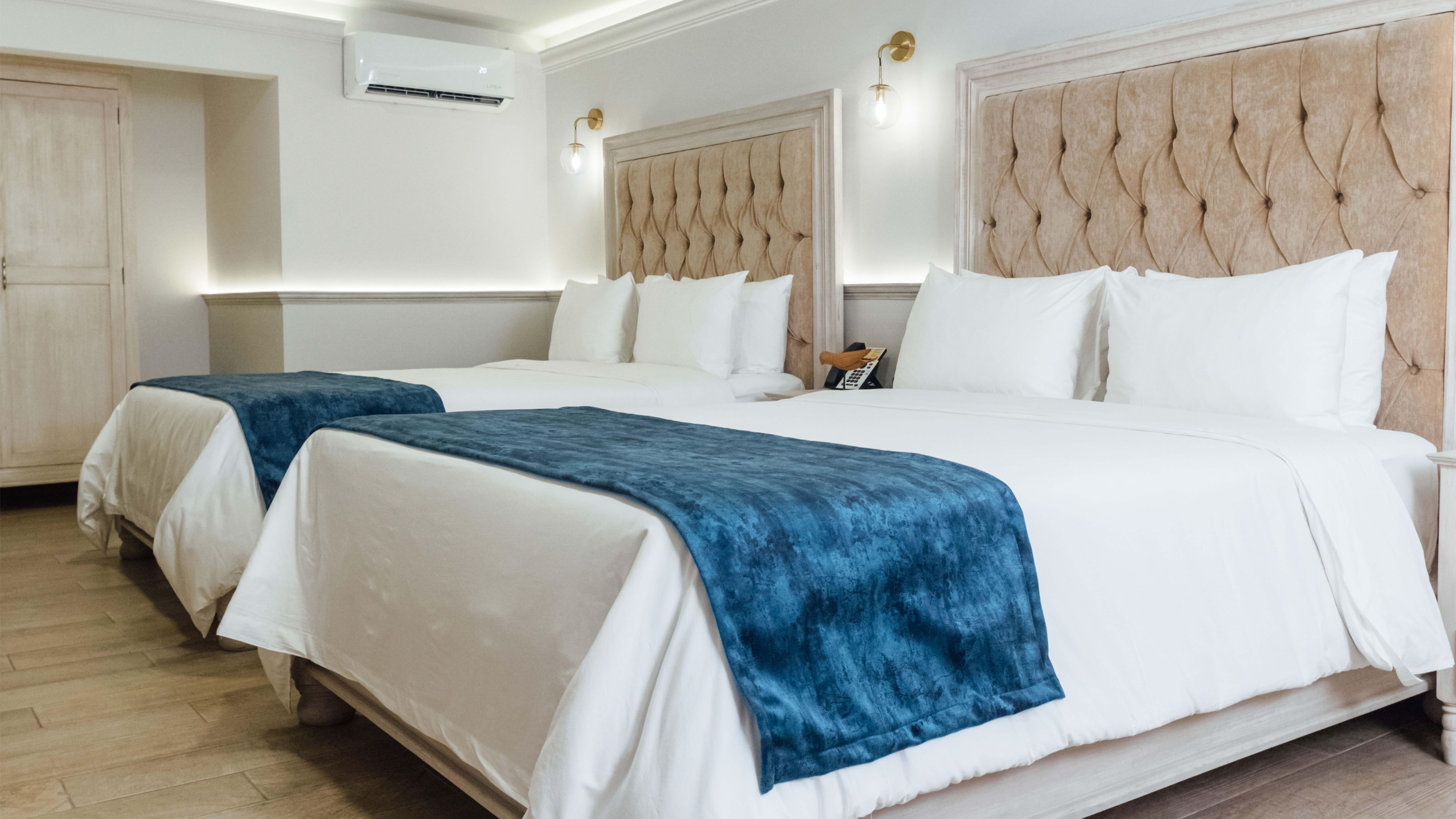 A hotel room with two beds, a large padded headboard, a wooden floor, and a wall-mounted air conditioner.