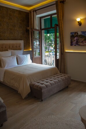 A bedroom with a large bed, a bench, a view through a window, and a painting on the wall. - Voco Royalty Puebla Downtown (Puebla)