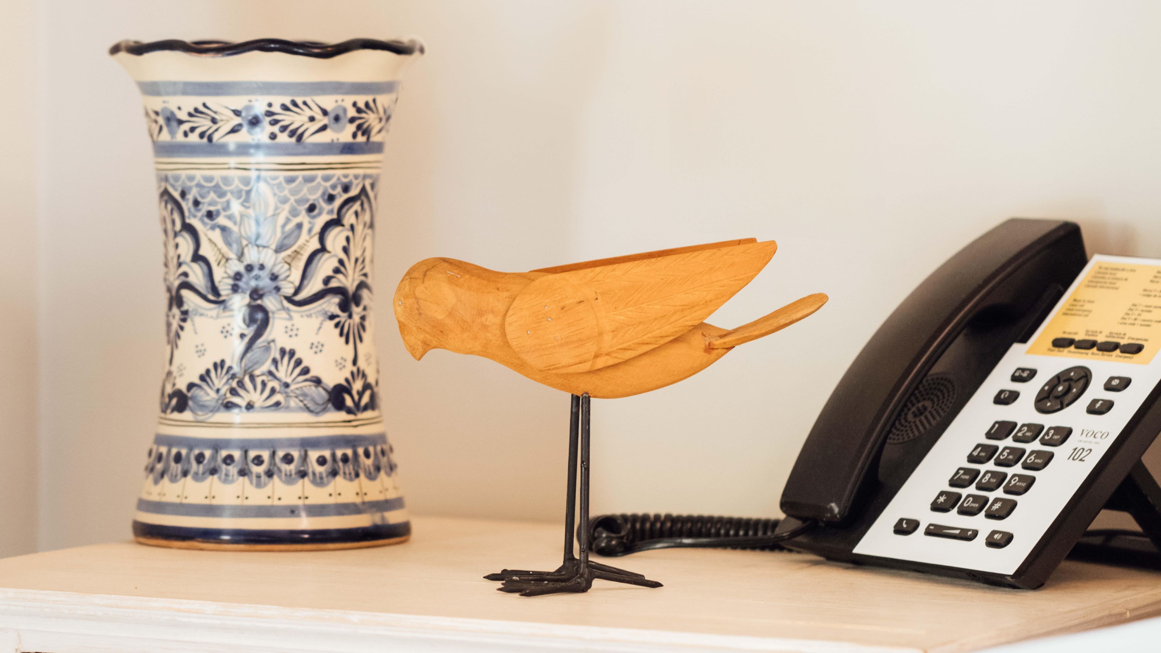 Un jarrón azul y blanco de cerámica, una escultura de pájaro de madera y un teléfono sobre una mesa.