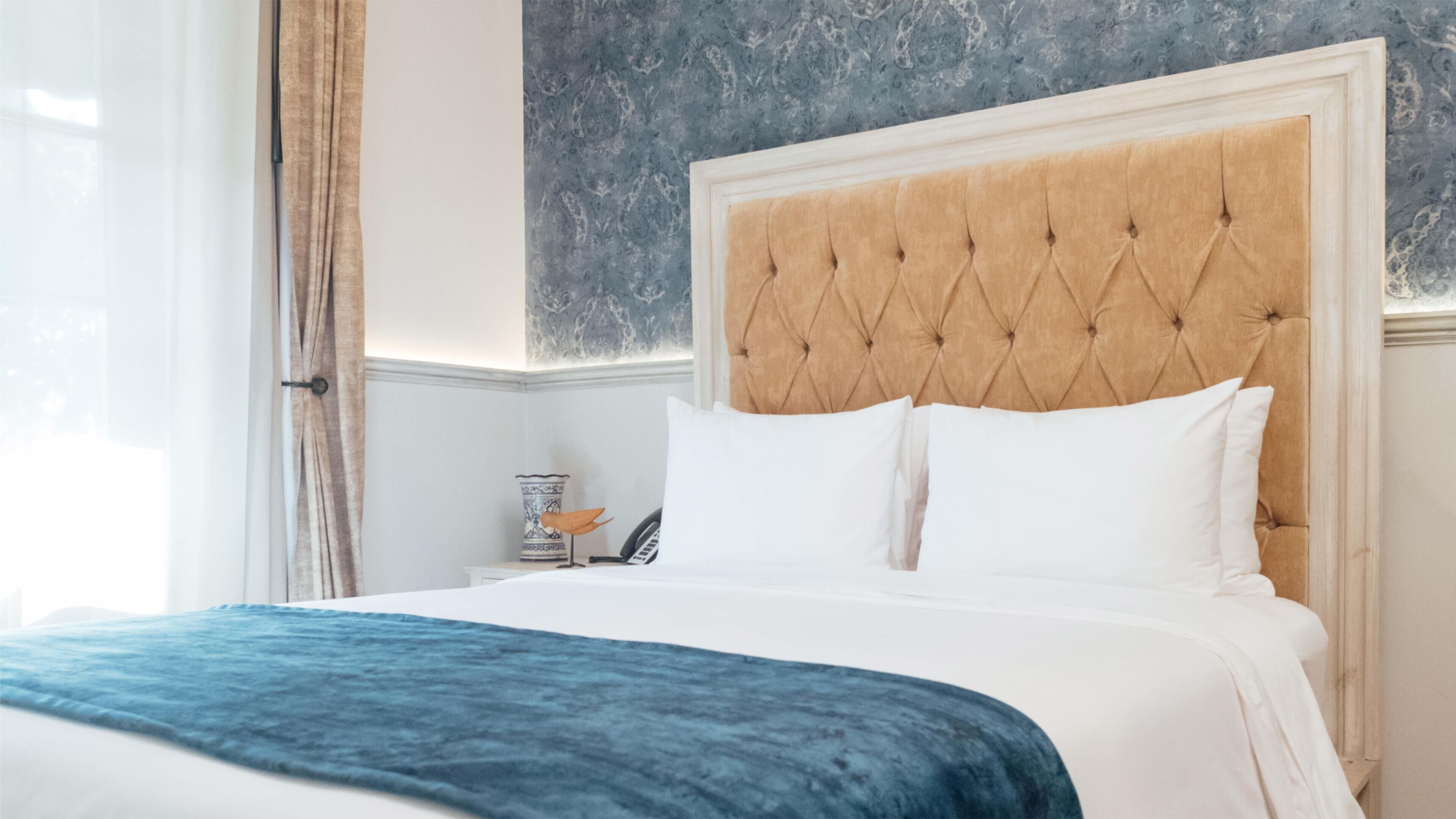 Une chambre d’hôtel avec un grand lit, une tête de lit capitonnée, du linge de lit blanc et un coussin décoratif bleu.