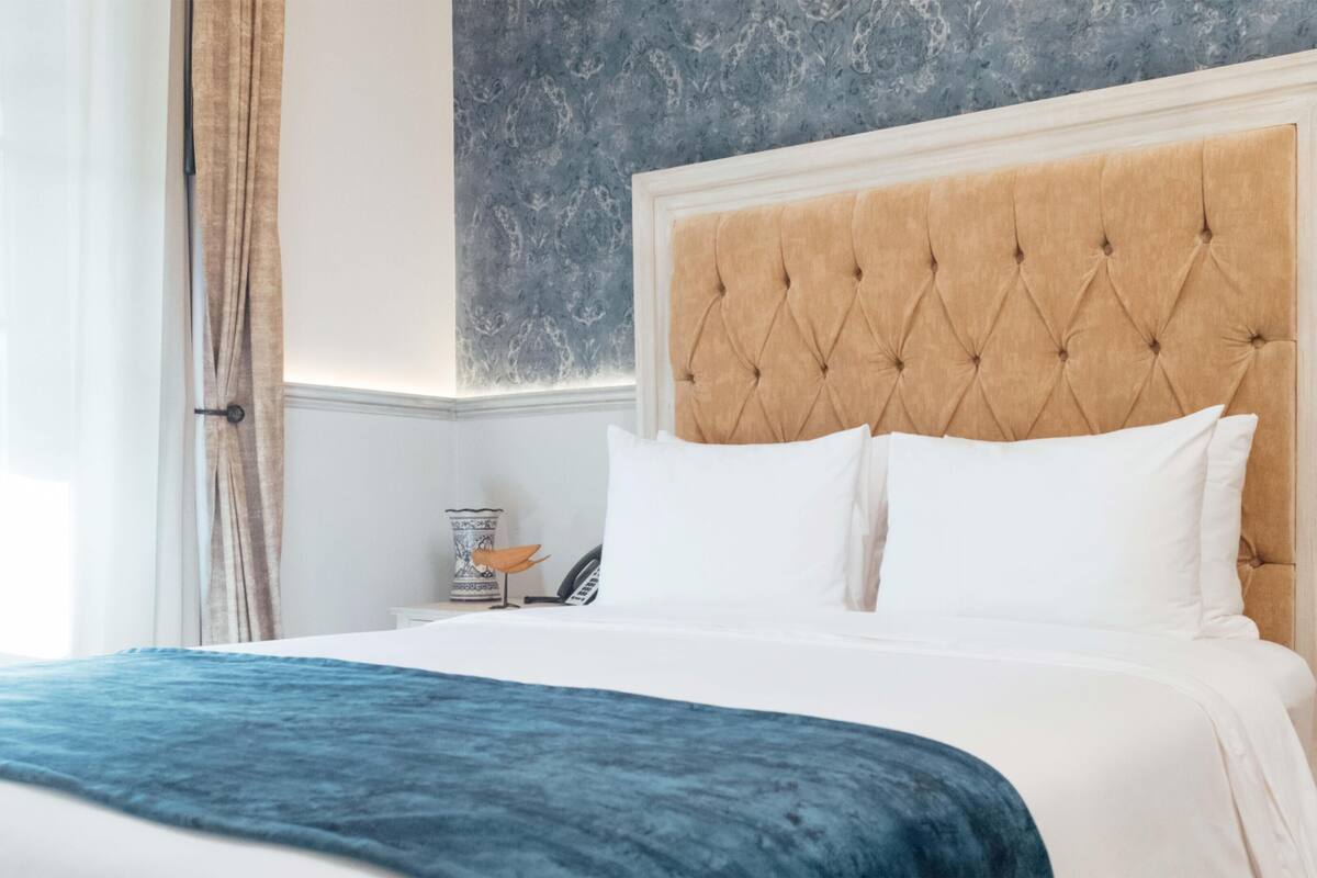 A hotel room with a large bed, a tufted headboard, white bedding, and a blue accent throw.
