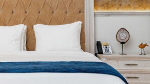 A neatly made bed with white linens and a blue accent, a wooden headboard, a bedside table with a telephone and a decorative item, and a wooden nightstand with drawers. - Voco Royalty Puebla Downtown (Puebla)