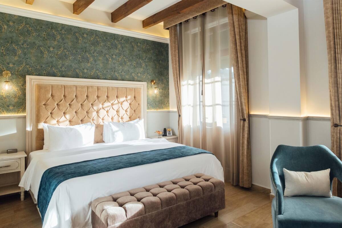 A bedroom with a large bed, a tufted headboard, a bench, a chair, and a nightstand with a lamp.