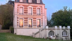 A large, multi-story building with a symmetrical facade, featuring a central entrance with a staircase, multiple windows with shutters, and a brick exterior. - Royal Rose (Étretat)