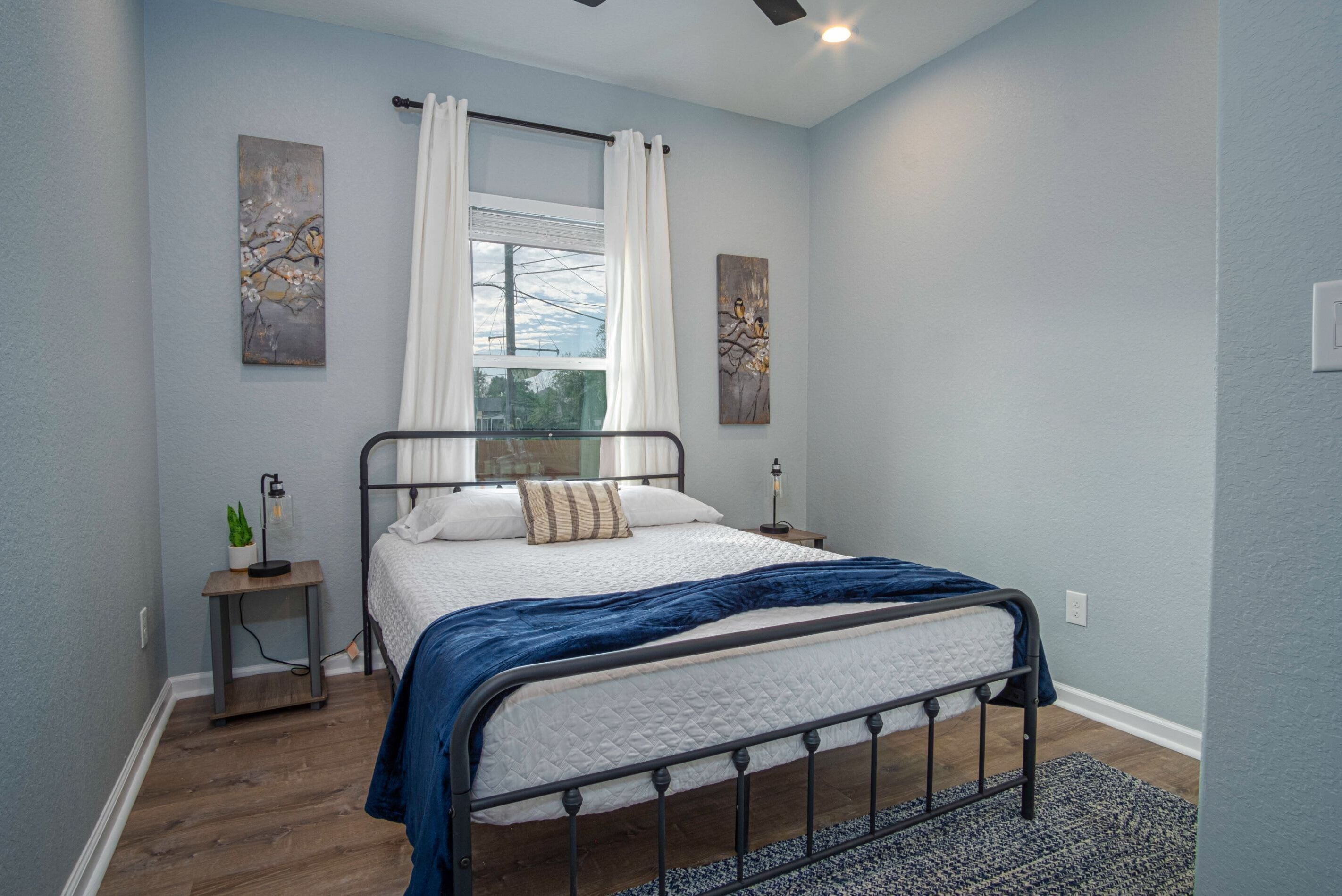 A bedroom with a bed, a nightstand, a lamp, a window with curtains, and artwork on the walls.