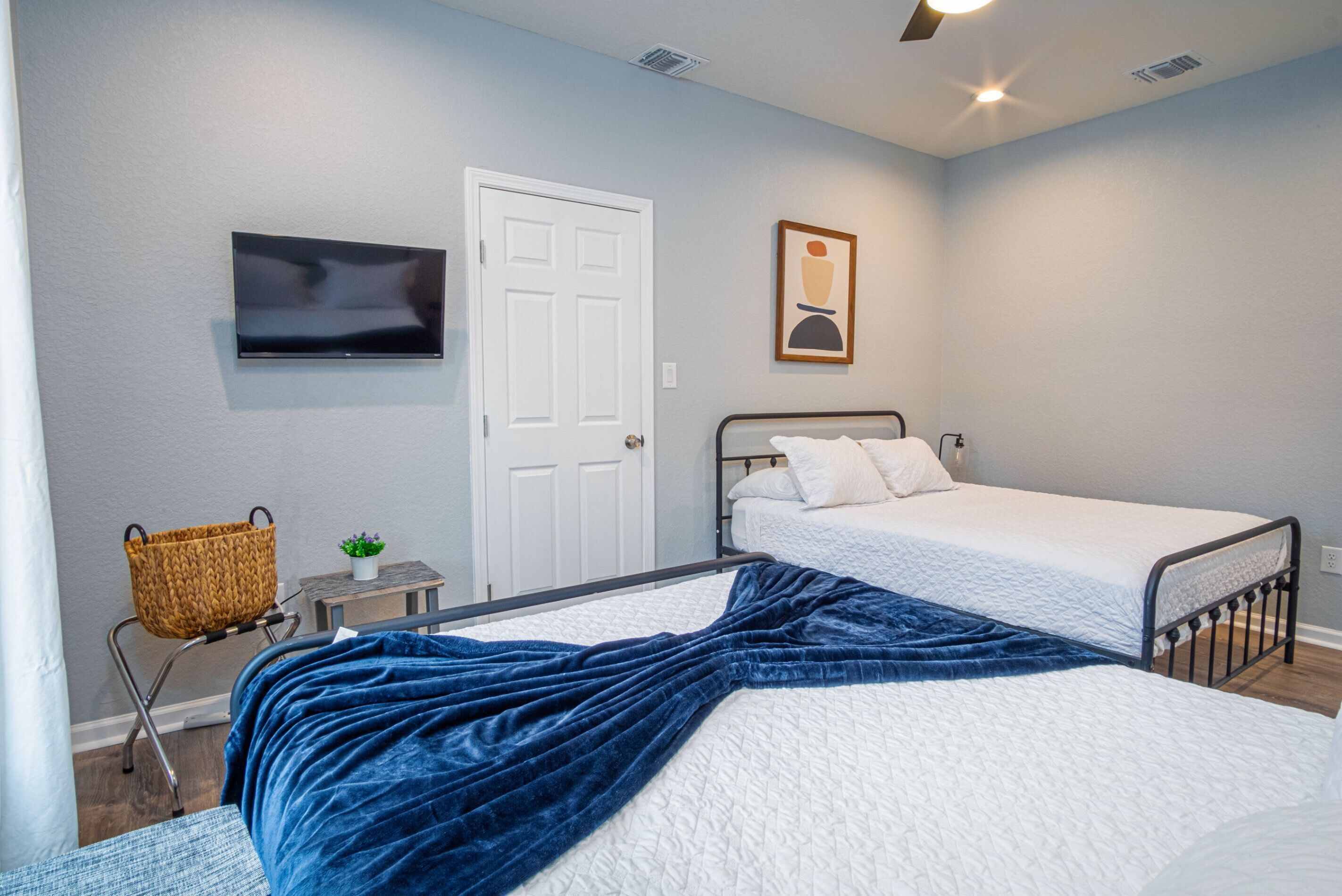 A bedroom with two single beds, a TV mounted on the wall, a door, and a small table with a basket and plant.