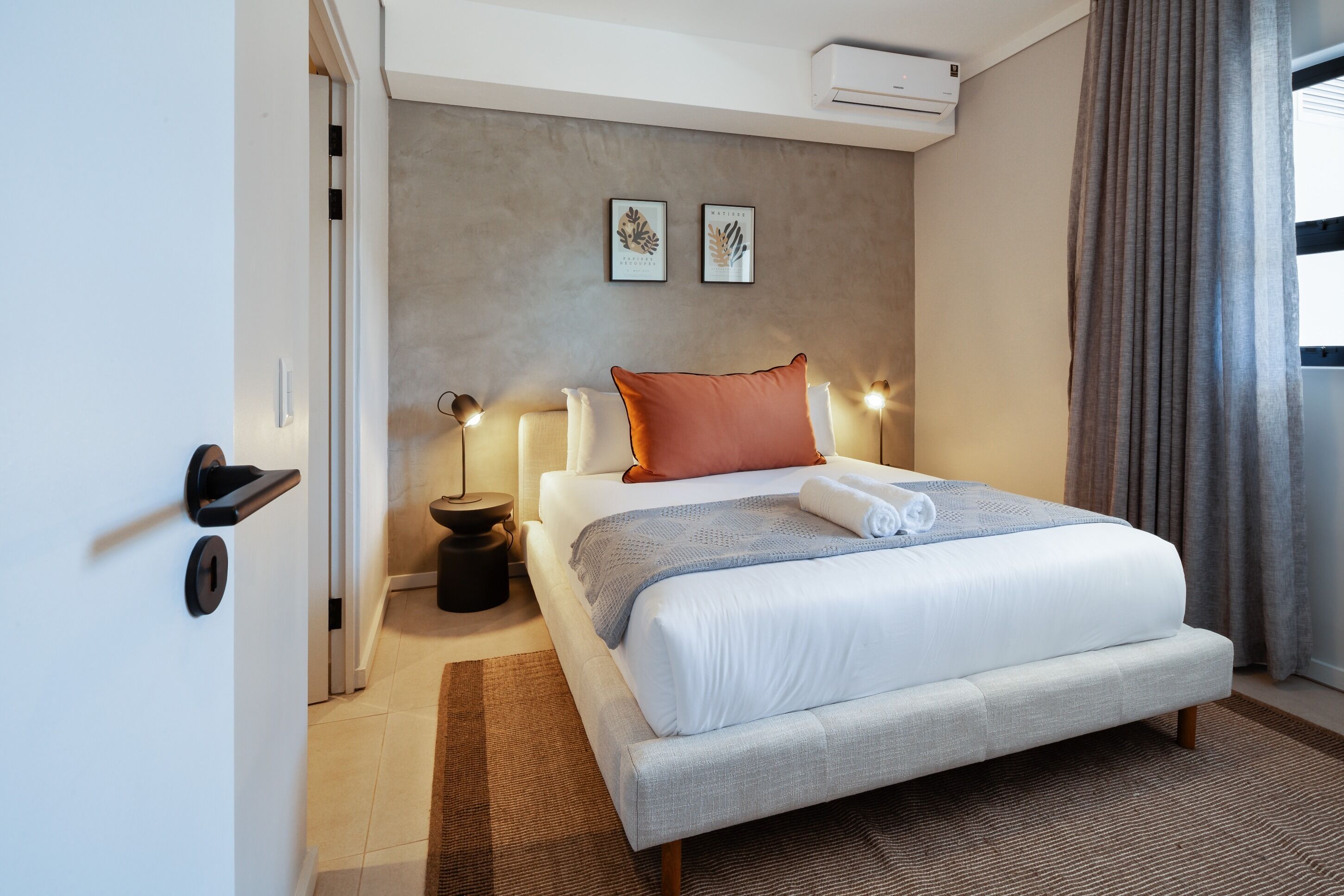A modern hotel room with a large bed, a bedside table with a lamp, and a wall-mounted air conditioner.