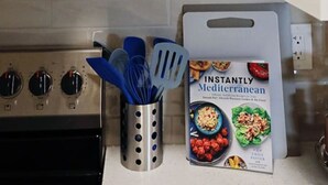 Eine Küche mit einer Edelstahl-Dunstesse, einem weißen Schrank mit Weingläsern und Einmachgläsern sowie einem Kochbuch mit dem Titel 'Instantly Mediterranean' auf der Arbeitsfläche.