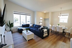 A modern living room with a dark blue sofa, white coffee table, and dining area with white chairs and a table. - The Quartz | Central 5BR | A/C | Garage | Expo (Edmonton)