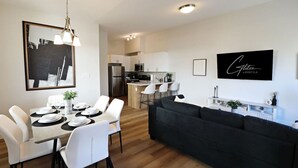 A modern living room with a dark sofa, a dining area with white chairs and a table set for four, and a kitchen in the background.