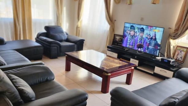 A living room with a large flat-screen TV, black leather sofas, a wooden coffee table, and beige curtains. - 3 Bed House - Sleeps 9 - Pets - Garden - Parking (Accra)