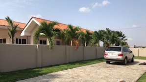 A residential area with a paved driveway, a white car parked, and a beige wall with palm trees. - 3 Bed House - Sleeps 9 - Pets - Garden - Parking (Accra)
