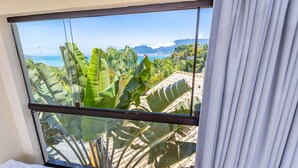 A room with a large window overlooking a tropical landscape with banana plants and a view of the sea and mountains. - Eco Baepi Residence (Ilhabela)