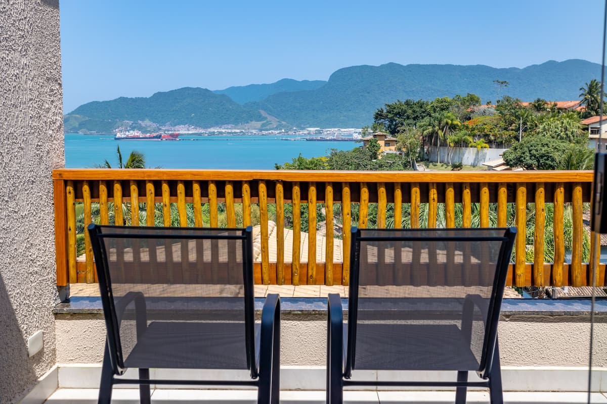 A balcony with two black mesh chairs, a wooden railing, and a view of a coastal area with mountains and buildings.