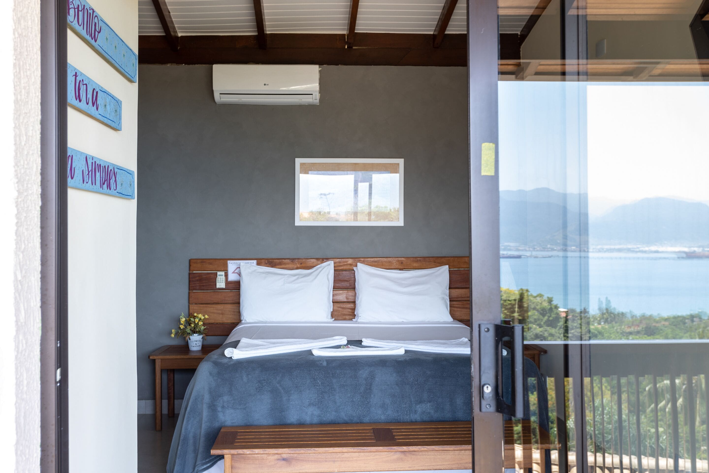 Quarto com cama, mesa de cabeceira, uma pequena mesa com planta, janela com vista e varanda com guarda-corpo.