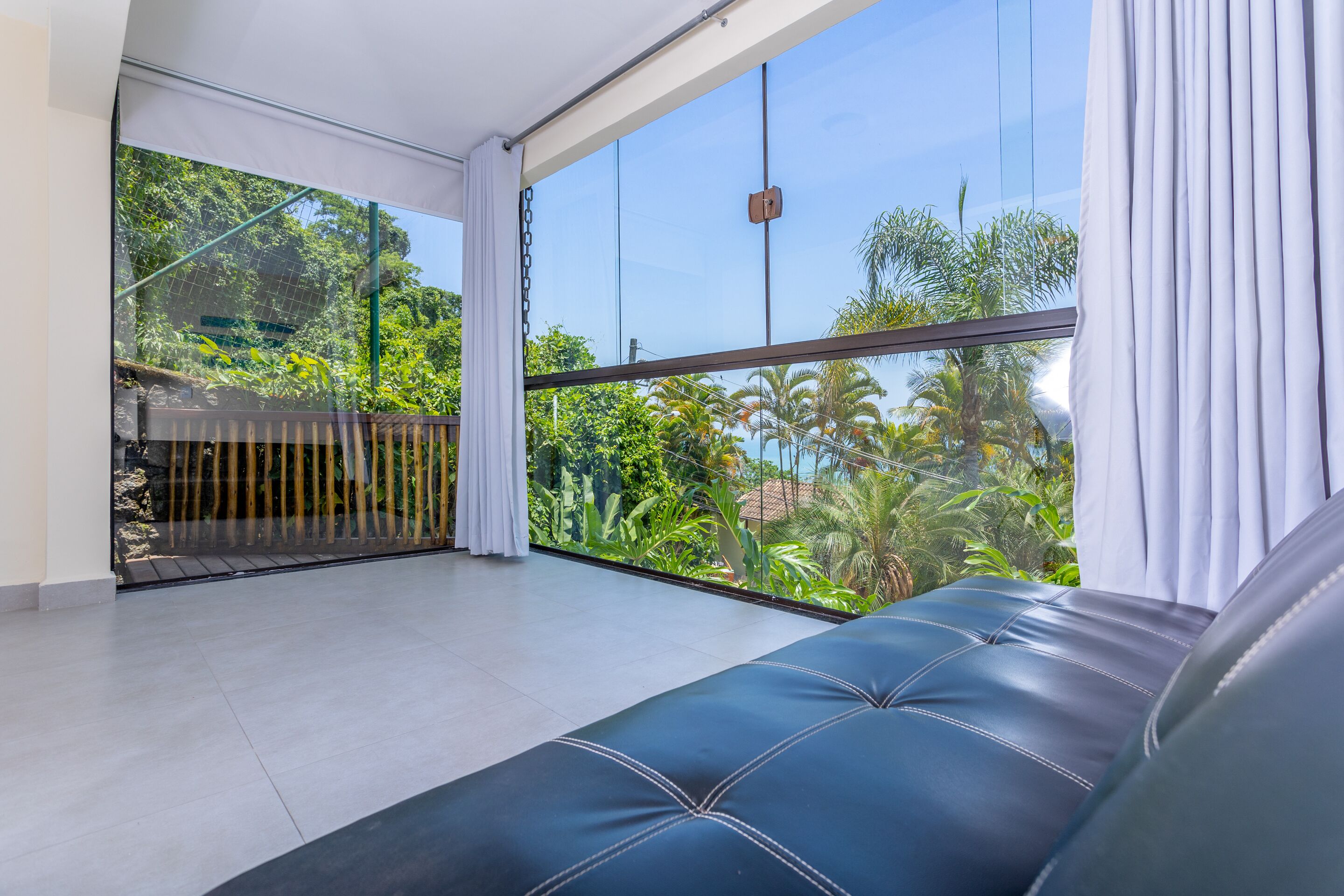 A modern room with a large glass window, a balcony with a view of greenery, and a black leather sofa.