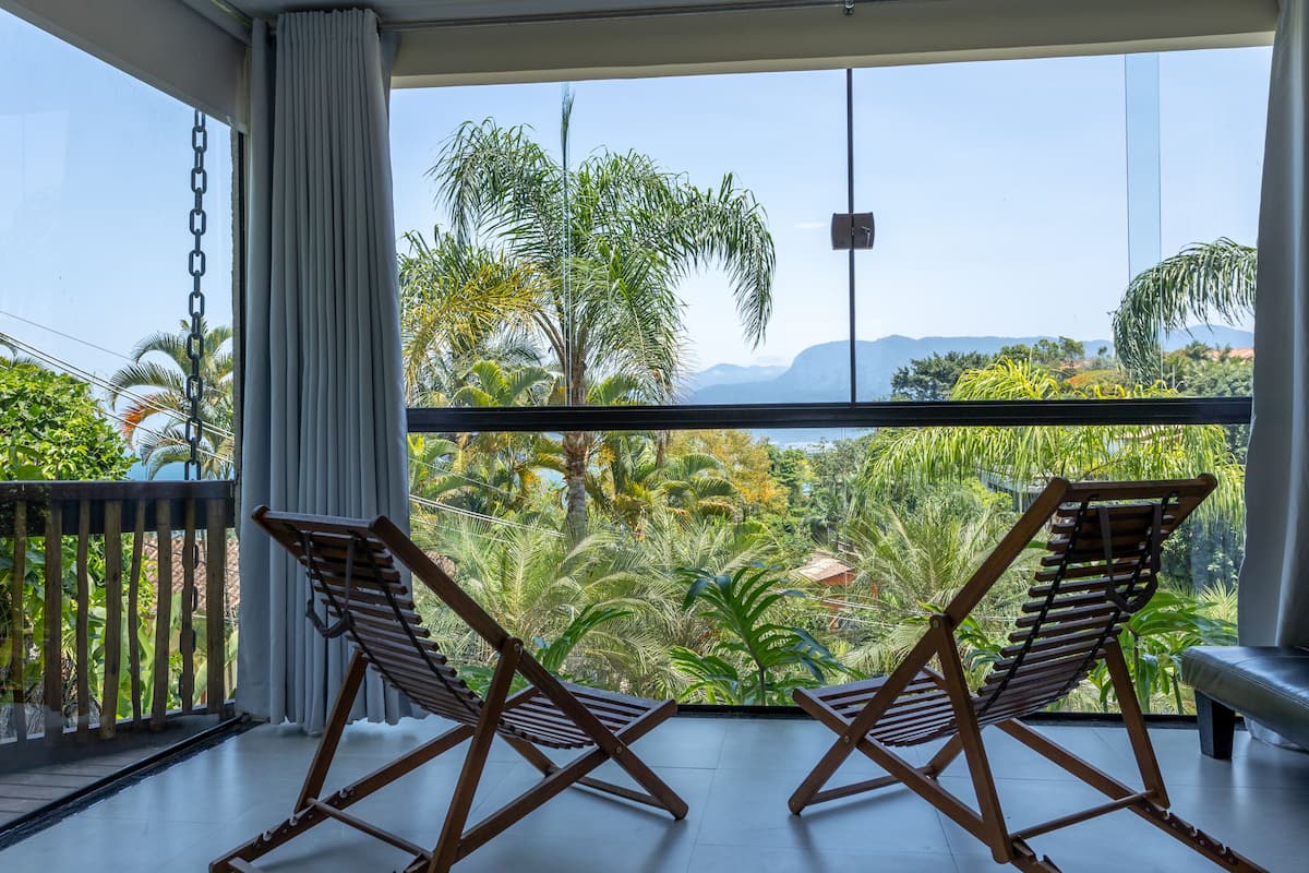 A balcony with two wooden chairs, a white curtain, and a view of tropical vegetation and mountains.