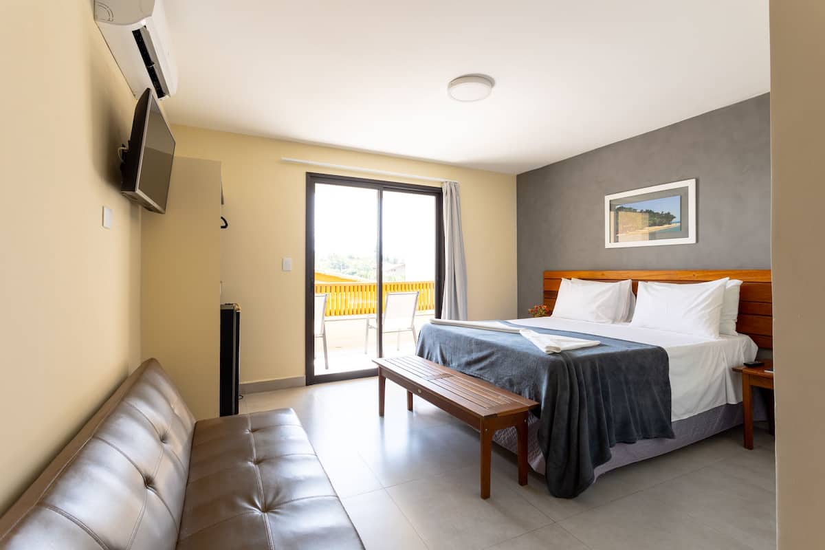 A hotel room with a bed, a bench, a TV, and a balcony with a view.