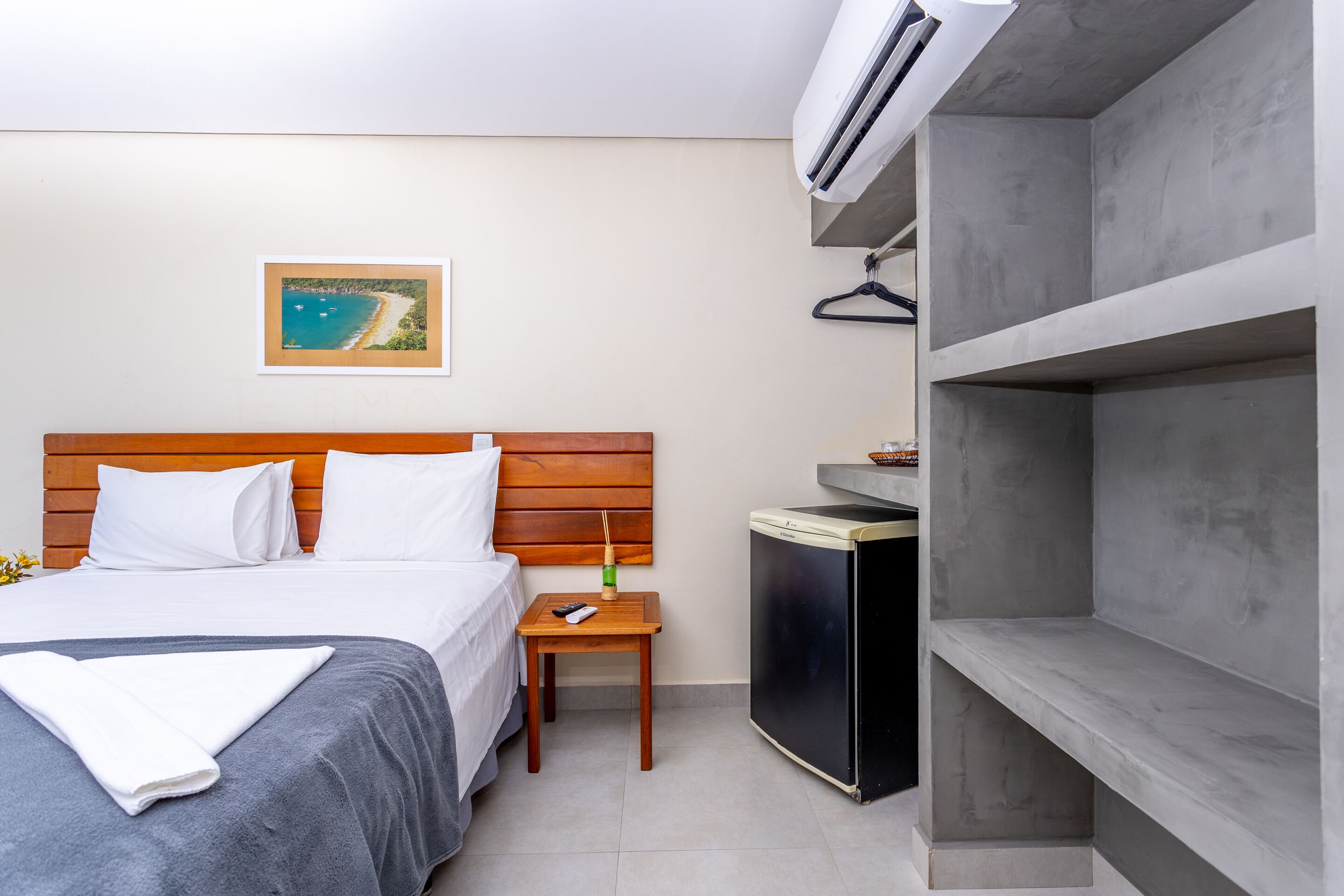 A hotel room with a bed, a small table with a remote control, a TV, and a wall-mounted air conditioner.