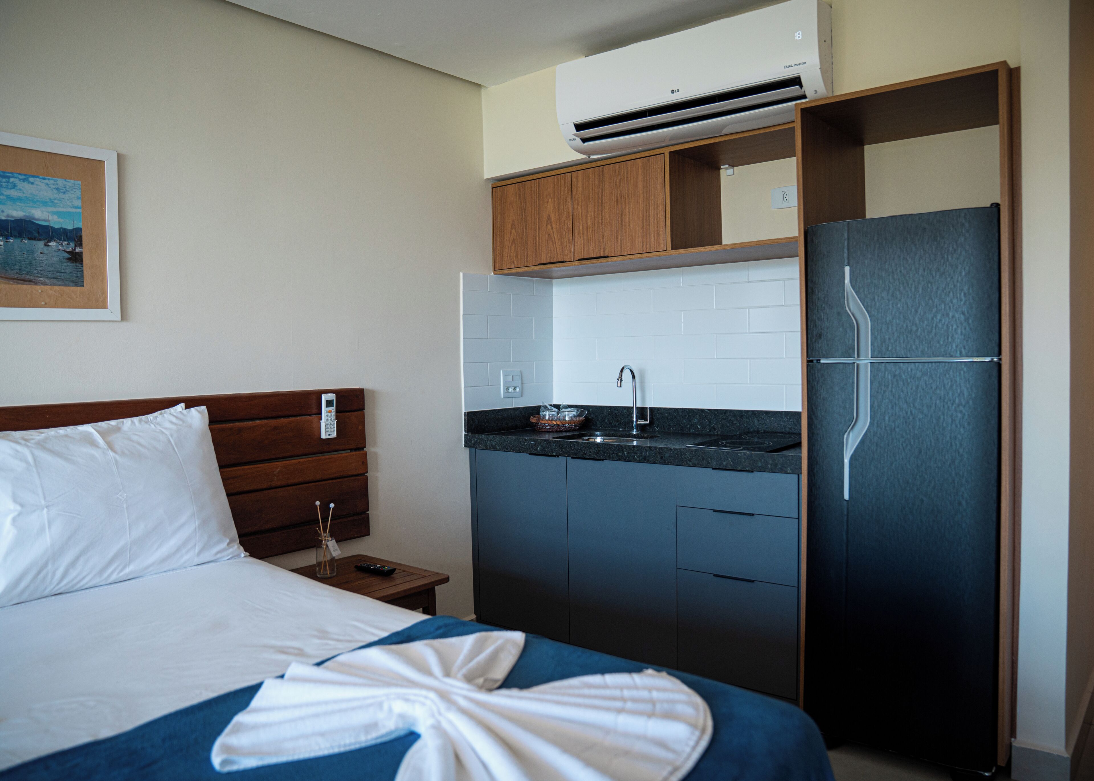 Habitación de hotel con cama, mesita de noche con control remoto, pequeña cocina con pileta y alacenas, y un refrigerador.