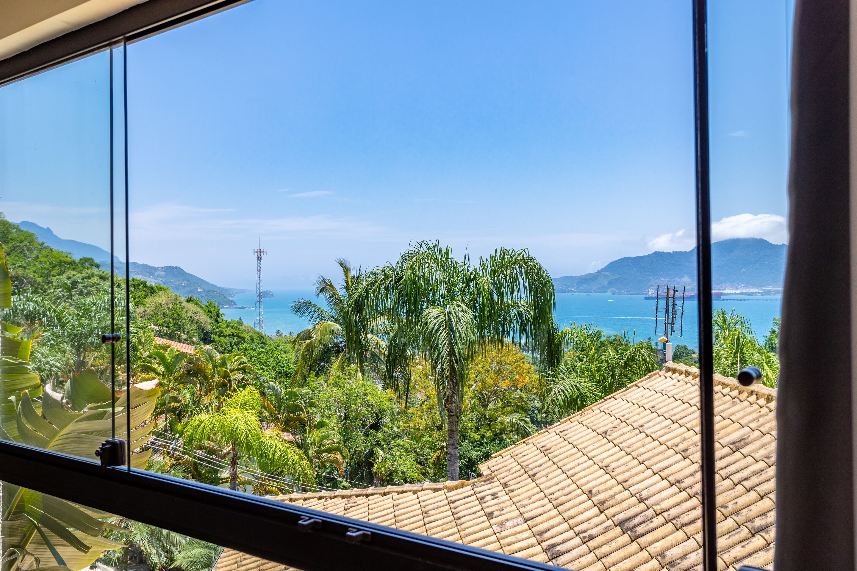 Une vue depuis une fenêtre donnant sur un paysage côtier, avec des palmiers et un toit recouvert de tuiles.