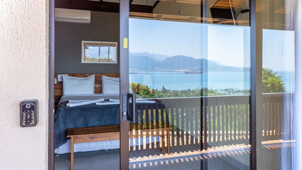 A bedroom with a bed, a bedside table, a window, and a view of the sea and mountains. - Eco Baepi Residence (Ilhabela)
