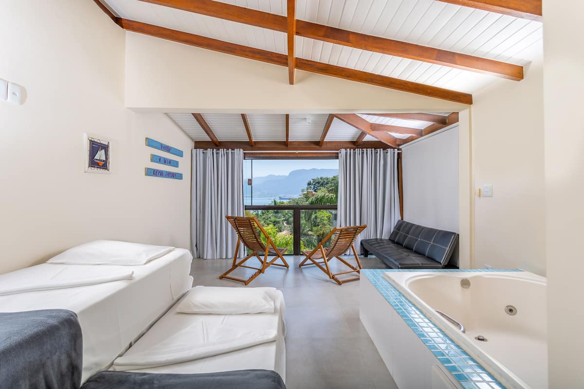 A room with a bed, a sofa, a small table, two chairs, a bathtub, and a view of mountains through large windows.