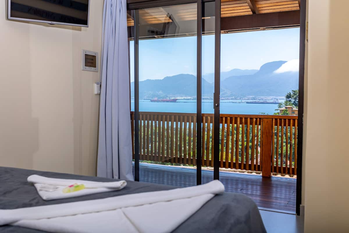 A hotel room with a bed, a view of a body of water, and a balcony with a wooden railing.