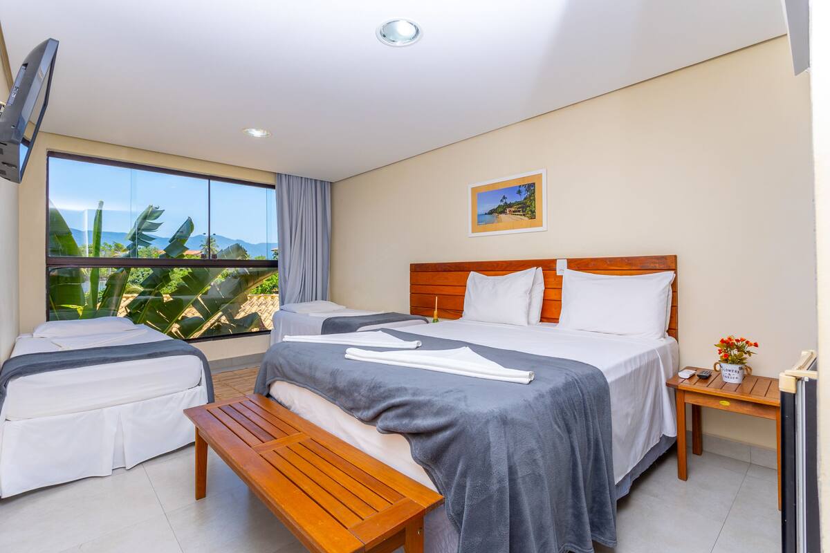 A hotel room with two beds, a wooden bench, a small table, a TV, and a large window with a view of greenery and mountains.