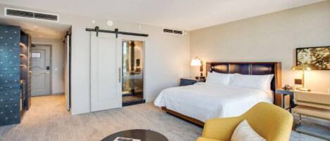 A hotel room with a bed, a yellow armchair, a bedside table with a lamp, and a door leading to a bathroom.