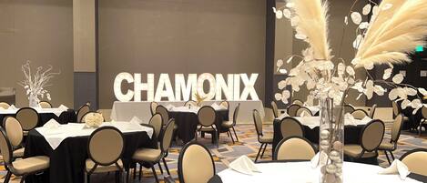 A banquet hall with tables set for dining, a large illuminated sign reading "CHAMONIX", and decorative elements like pampas grass and a vase with ornaments.