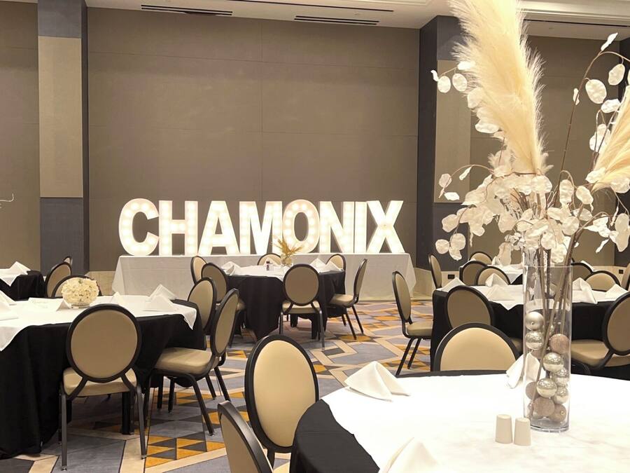 En festlokale med dækkede borde, et stort oplyst skilt med teksten "CHAMONIX" og dekorative elementer som pampasgræs og en vase med pynt.