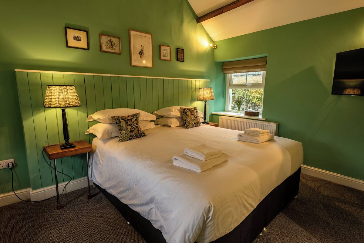 A neatly made bed with white linens and a set of towels, a bedside table with a lamp, and a window with a view of greenery.