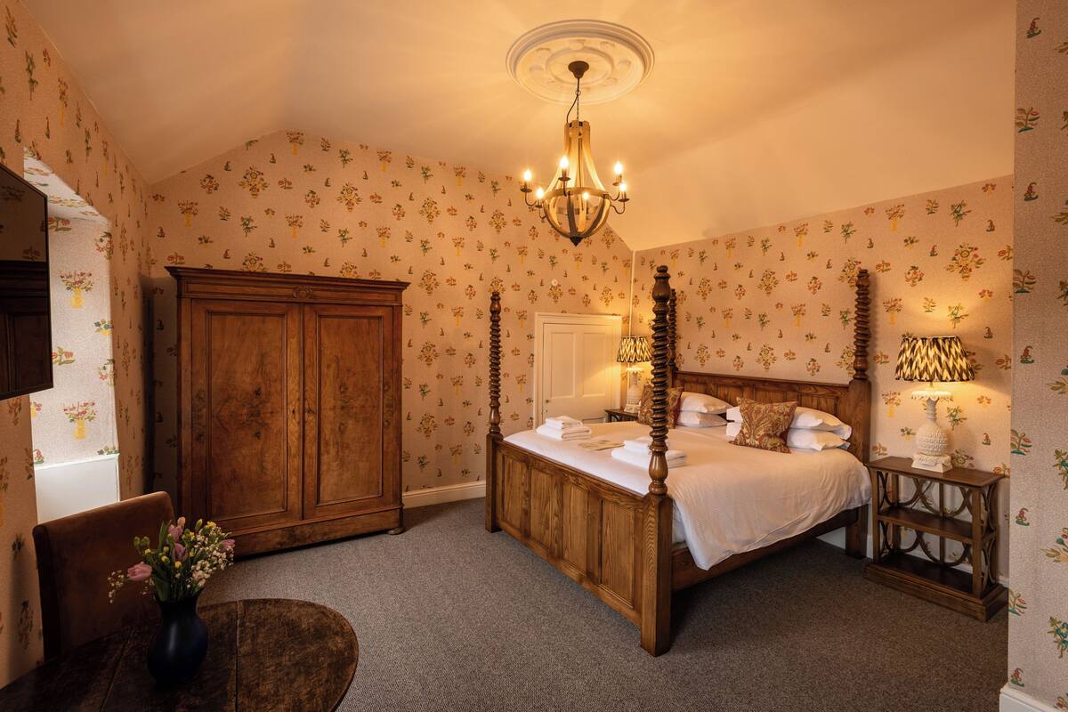 A traditional bedroom with a four-poster bed, wooden wardrobe, and floral wallpaper.