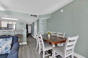 Uma área de refeições com mesa de madeira e cadeiras brancas, sofá azul e cozinha ao fundo.