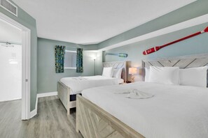 Quarto de hotel com duas camas, uma cabeceira de madeira, uma mesinha de cabeceira com luminária e uma janela com cortinas estampadas.
