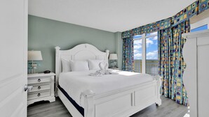 A neatly made bed with white linens and a decorative towel, a nightstand with a lamp, and a window with patterned curtains. - Baywatch Resort Incredible Oceanfront Family Dream Maker (North Myrtle Beach)