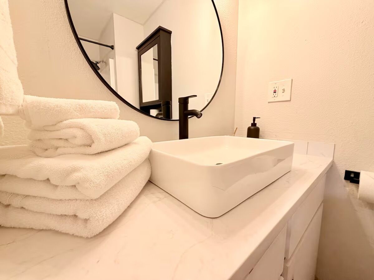 Casa de banho com lavatório branco, espelho e uma pilha de toalhas brancas.